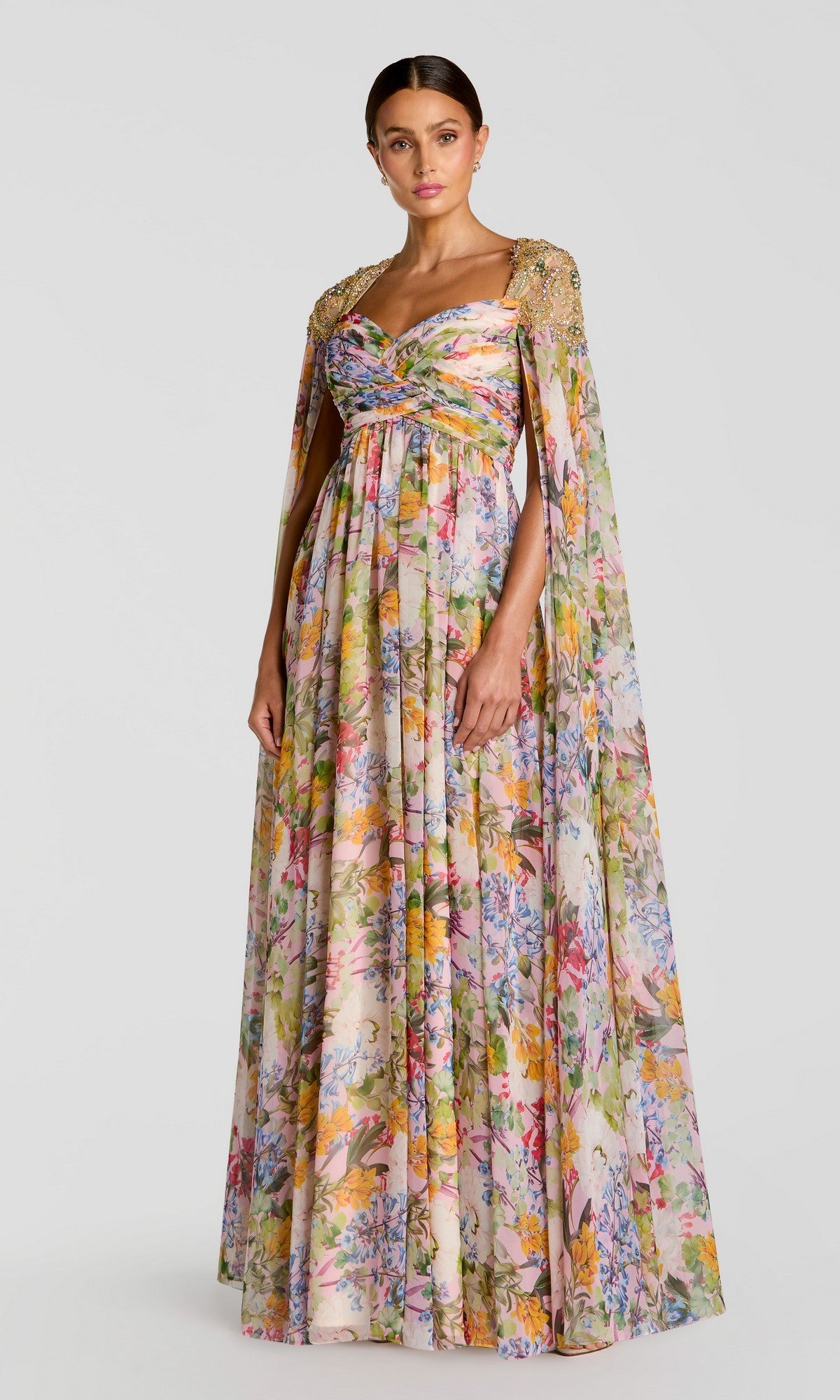 A woman wears the Mac Duggal 11990 long formal dress in pastel multi—a floor-length floral chiffon gown with a gathered bodice and attached cape with embellished shoulders—ideal for special occasions.